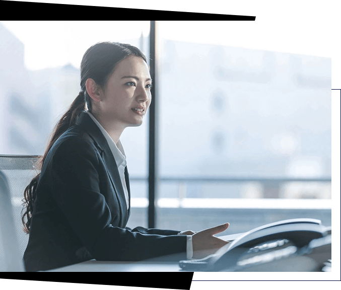 A woman in a business suit sits at a desk, gesturing while discussing recruiting services, with large windows and a blurred cityscape in the background.