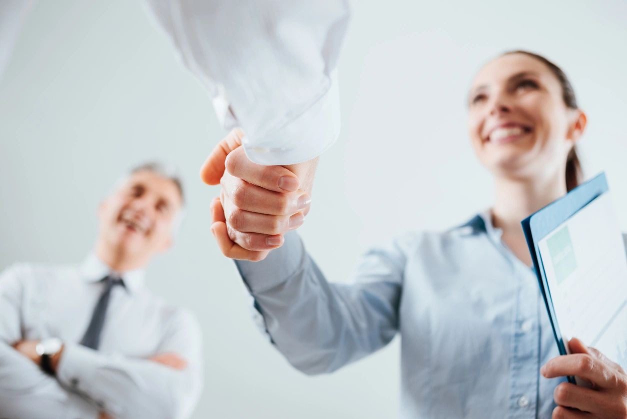 Two people shaking hands while another person stands in the background, smiling. One of the individuals holding a clipboard, indicating recruiting services at work.