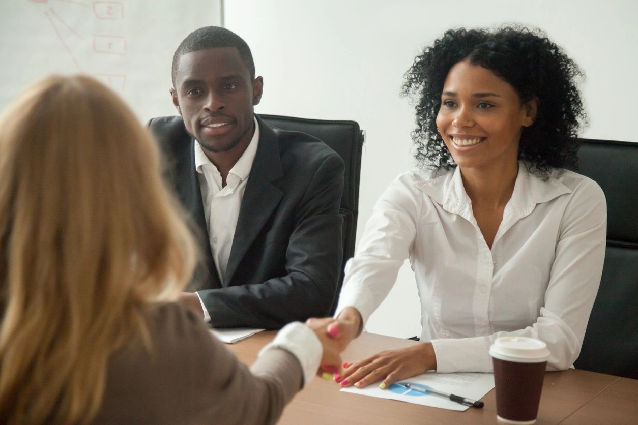 Two professionals sit at a table during a meeting, with one of them extending a handshake to a third person whose back is facing the camera. A cup of coffee and documents about recruiting services are on the table.